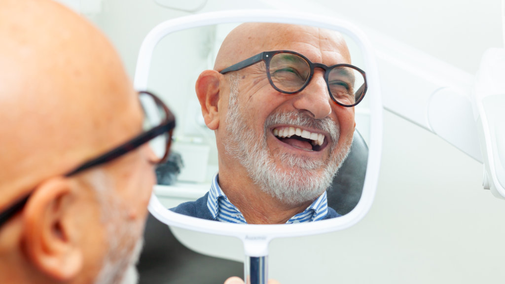 Senior male patient smiling while looking in mirror after getting dentures